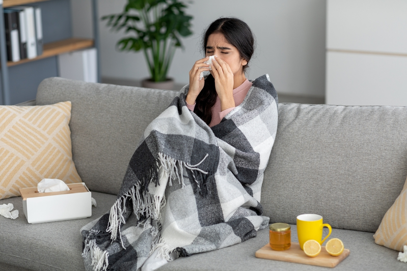 young woman wrapped up in blanket on couch blowing nose with a tissue
